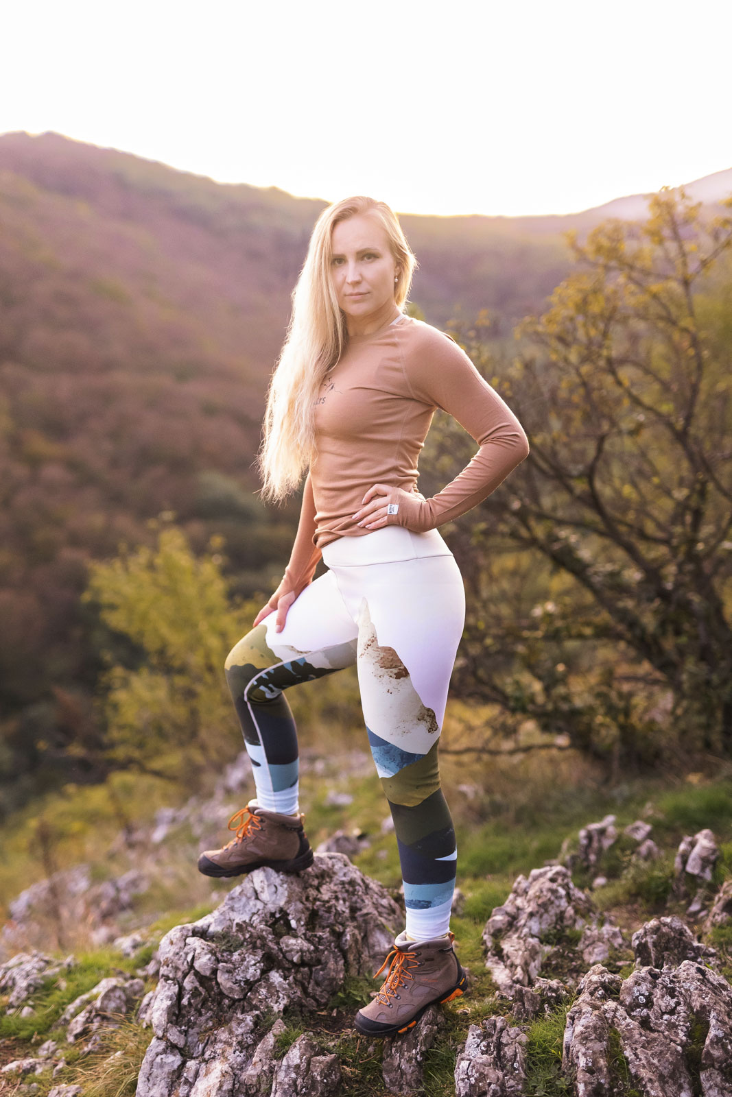 Žena stojí na kopci pri západe slnka. Má na sebe hnedé merino tričko s dlhým rukávom značky kallys.
