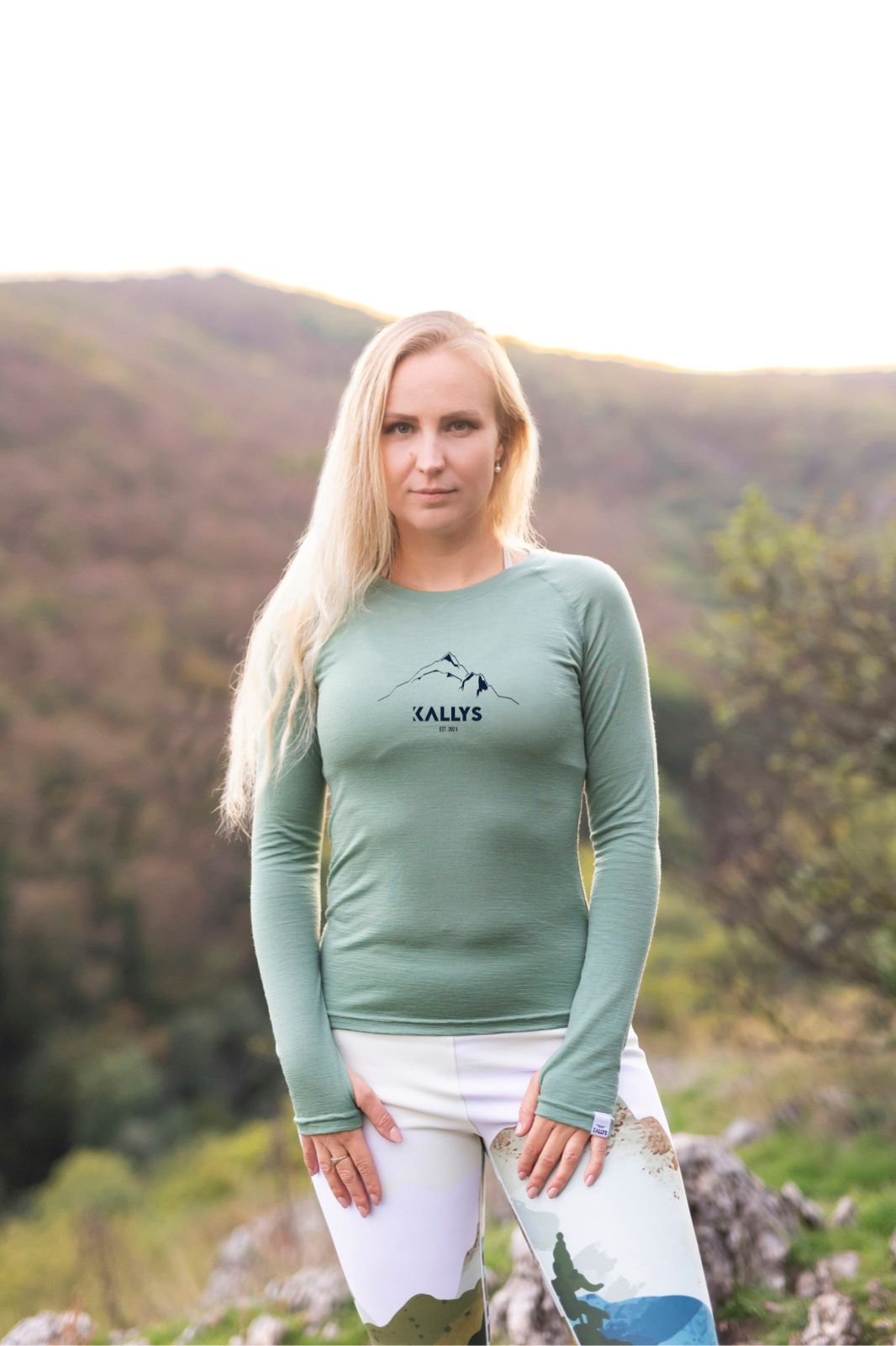 Blondínka stojí na vrchole kopca a má na sebe oblečené mentolovo zelené merino tričko s dlhým rukávom značky kallys. Tričko sa volá Summit.